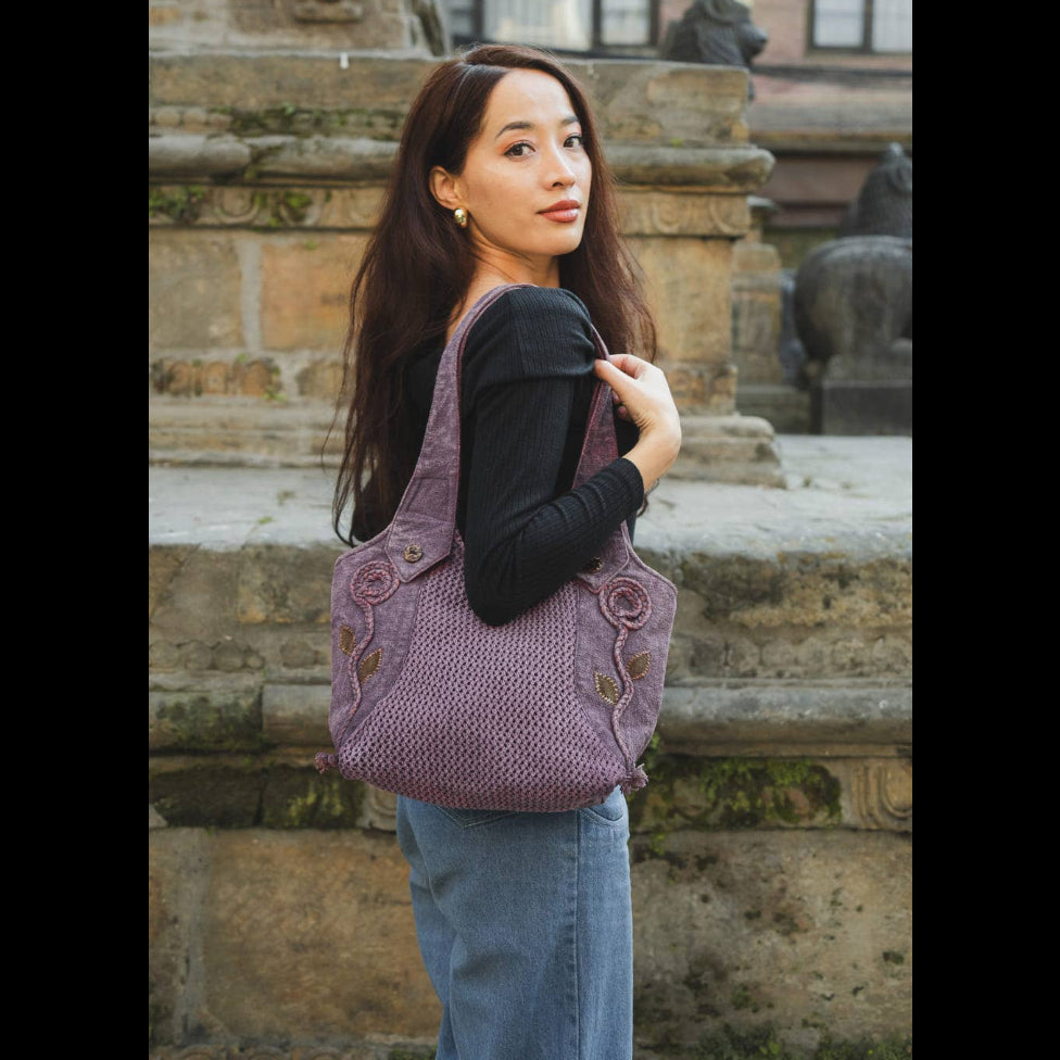 A woman wearing a black long-sleeve top and blue jeans carries a large, textured lavender-purple woven shoulder bag with decorative side details, standing in front of a stone architectural background.